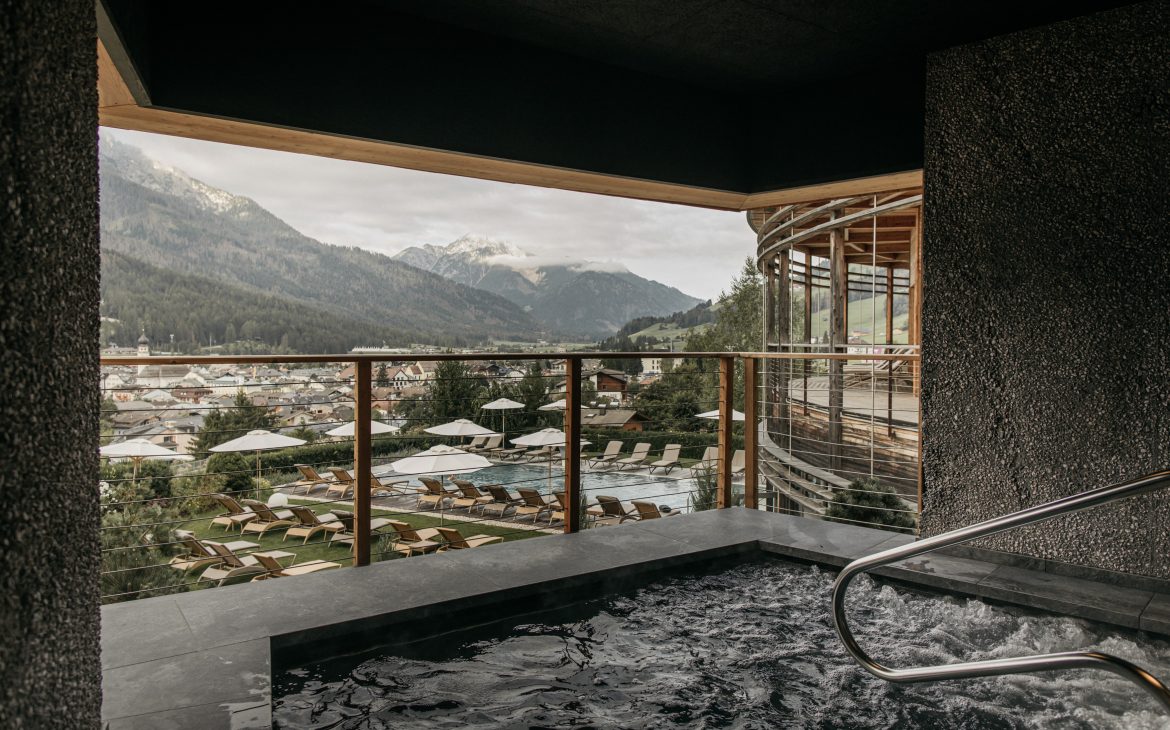 Im Vordergrund ein sechseckiger Pool mit sprudelndem Wasser, umrandet von dunklen Steinplatten. Im Hintergrund die Aussicht des Naturhotels Leitlhof auf Dorf und Berge.